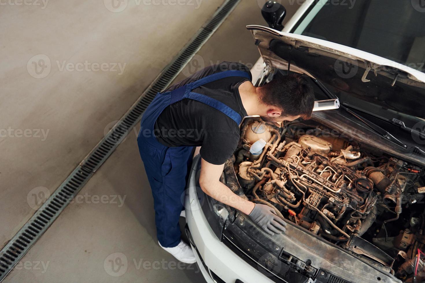 unter der Haube. Mann in Arbeitsuniform repariert weißes Auto drinnen. Konzept des Autoservices foto