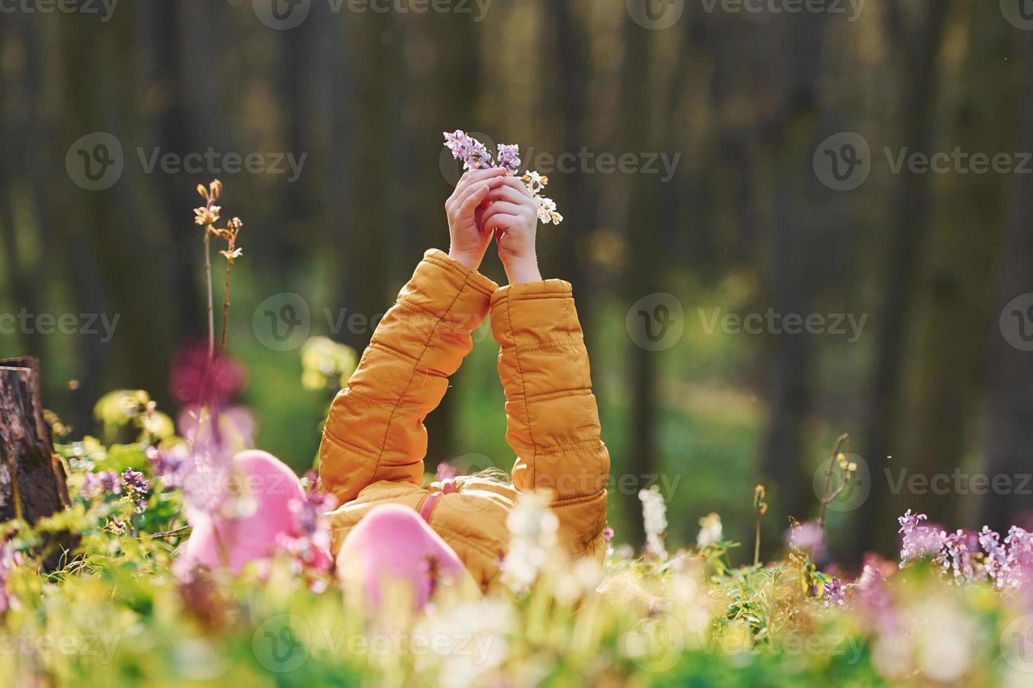 glückliches kleines Mädchen in Freizeitkleidung, das sich tagsüber im Frühlingswald auf den Boden legt foto