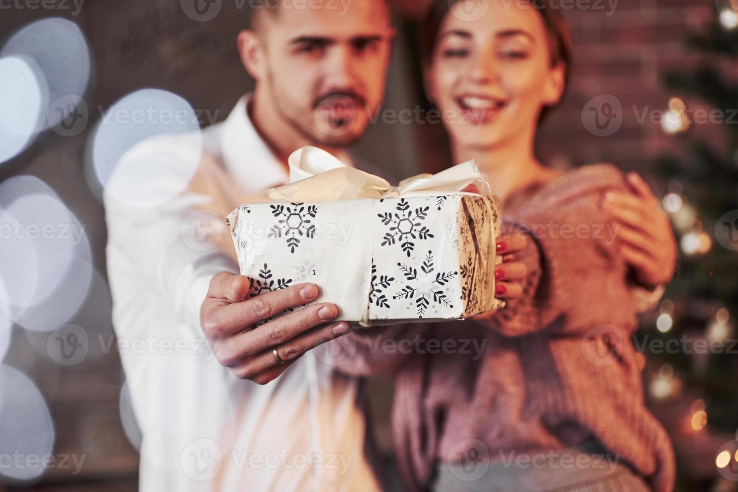 weiße Geschenkbox vor der Kamera halten. nettes paar feiert neujahr im neujahr dekorierten raum foto