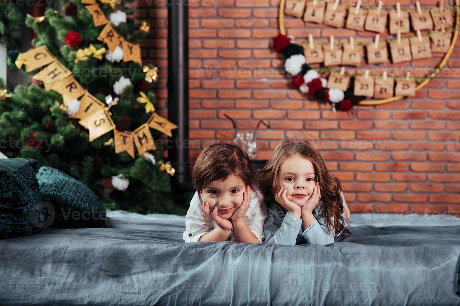 Zwei fröhliche weibliche Kinder liegen auf dem Bett mit Neujahrsdekorationen und Feiertagsbaum foto