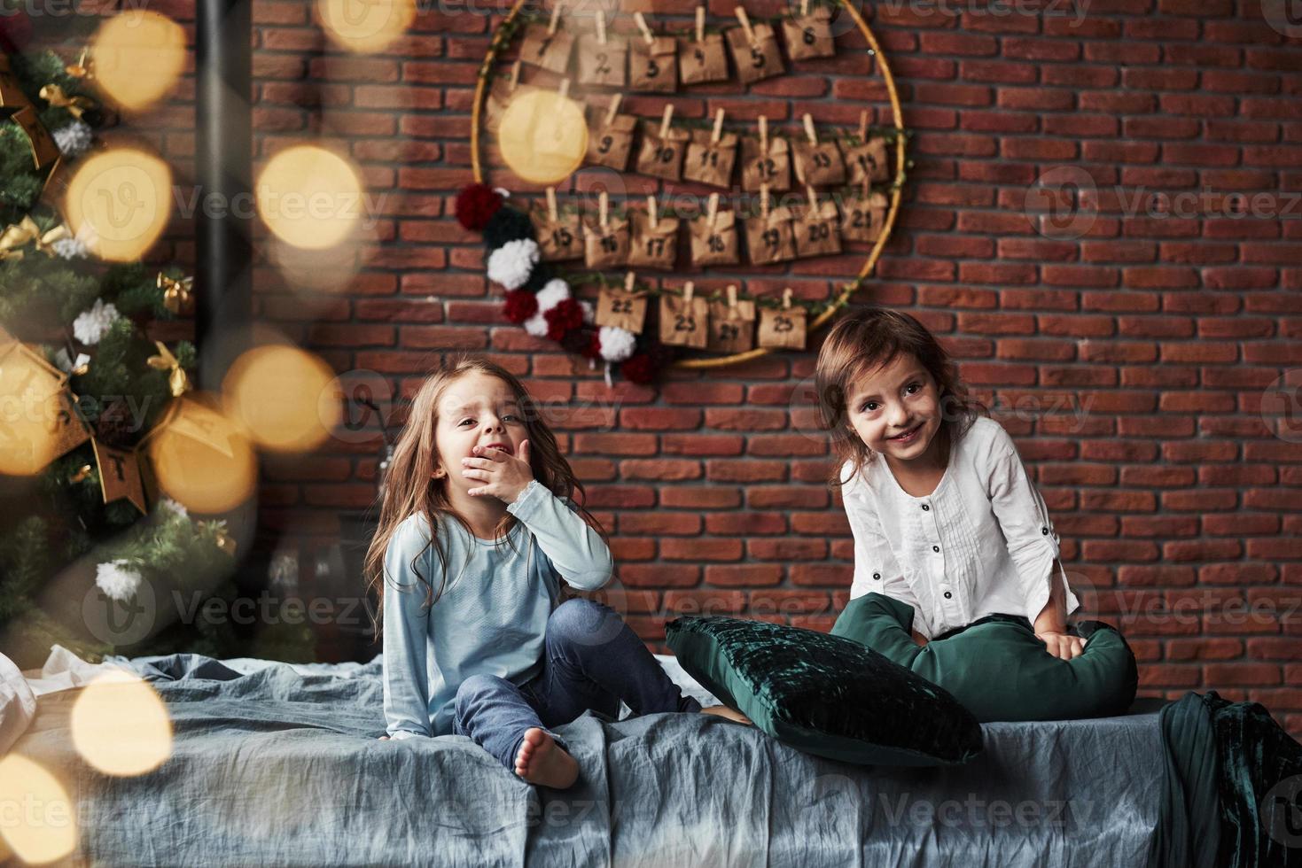 Weihnachtsbaum im Hintergrund schafft Neujahrsstimmung. Kleine Mädchen, die sich auf dem Bett mit Urlaubsinterieur im Hintergrund amüsieren foto