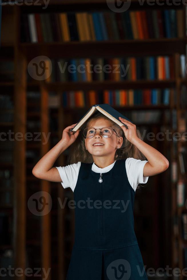 süßes kleines Mädchen mit Brille steht in der Bibliothek voller Bücher. Konzeption von Bildung foto