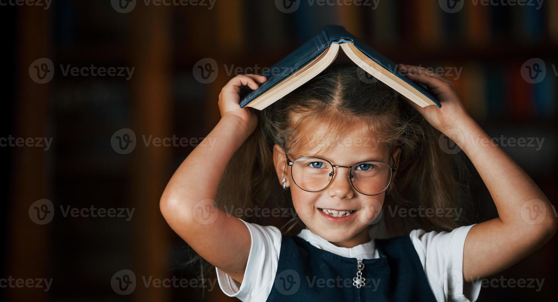 süßes kleines Mädchen mit Brille steht in der Bibliothek voller Bücher. Konzeption von Bildung foto