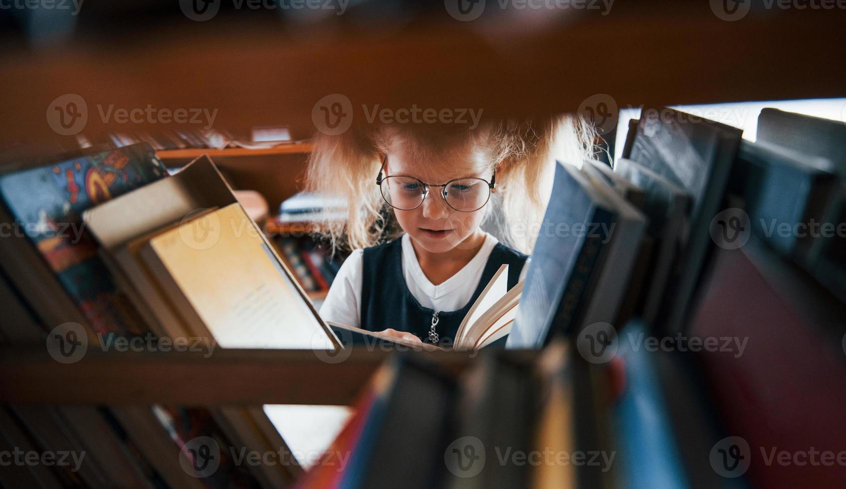 kleines Mädchen mit Brille, das in der Bibliothek nach einem Buch sucht. Konzeption von Bildung foto