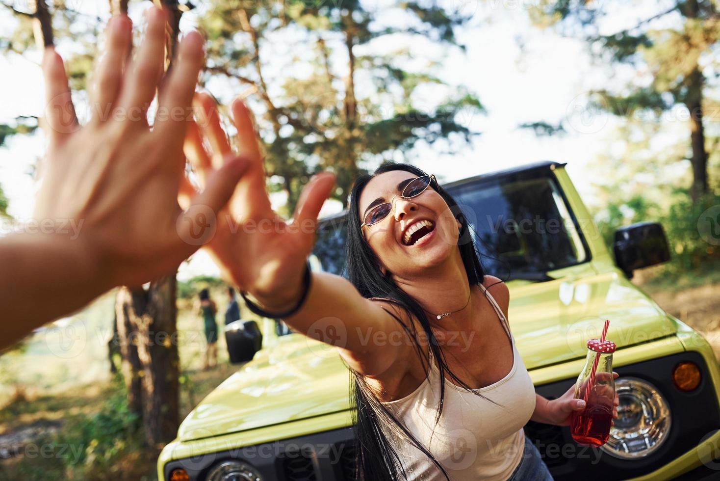 gibt High Five. blondine mit alkohol in der hand spazieren im wald gegen grünen jeep foto