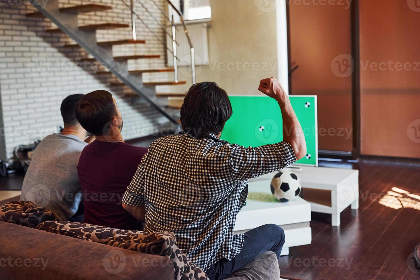 mit erhobenen Händen. aufgeregt drei freunde, die zu hause gemeinsam fußball im fernsehen gucken foto
