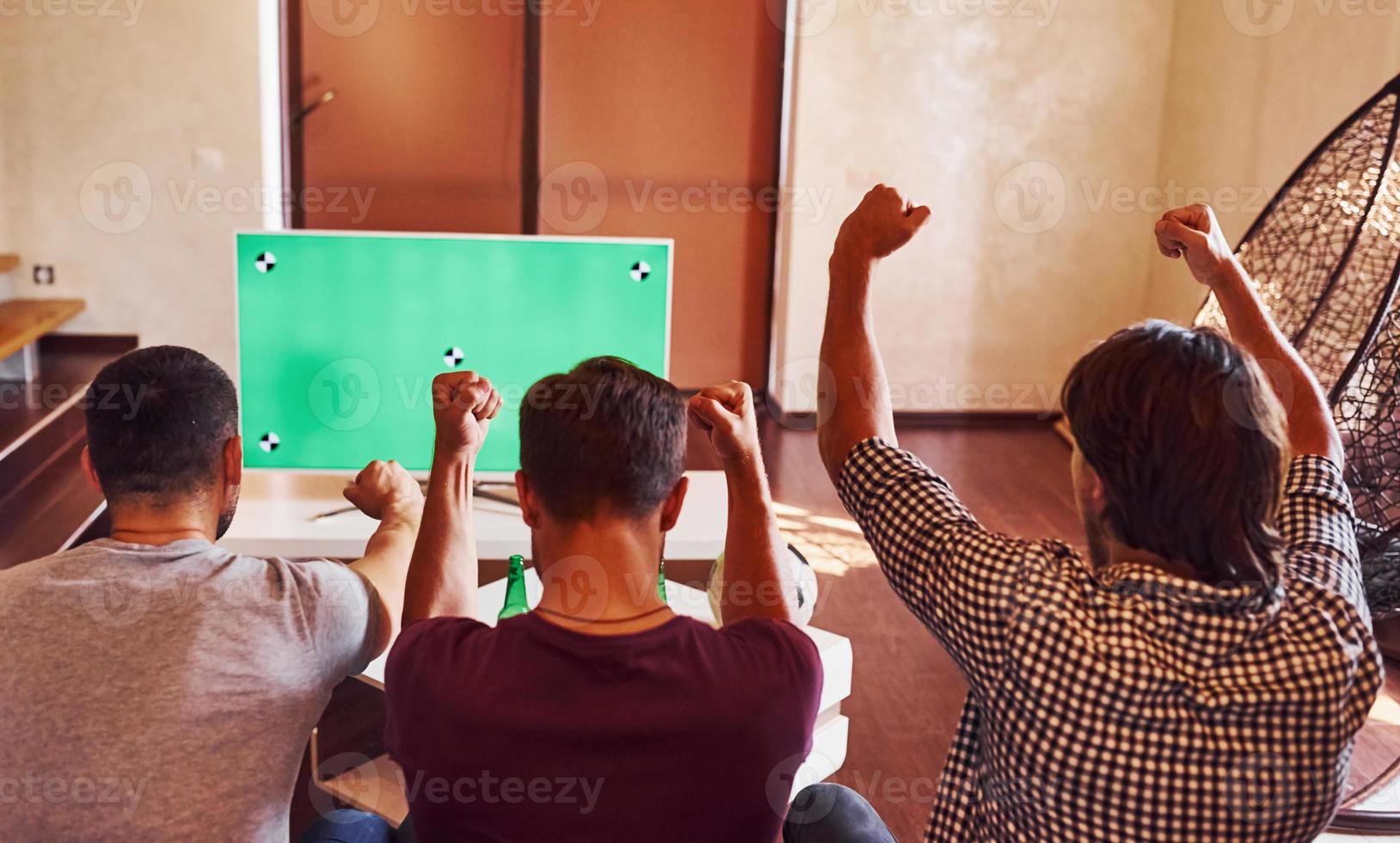 mit erhobenen Händen. aufgeregt drei freunde, die zu hause gemeinsam fußball im fernsehen gucken foto