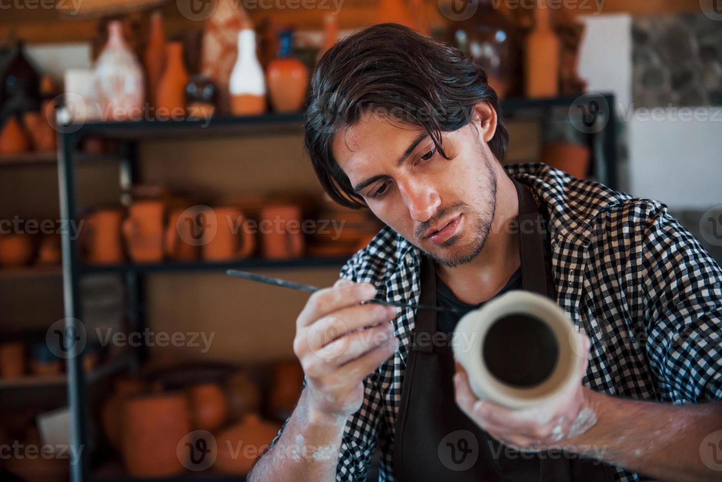 Der junge Keramiker hält seinen frischen handgefertigten Topf und macht mit einem Spezialwerkzeugstock etwas Detaillierung foto