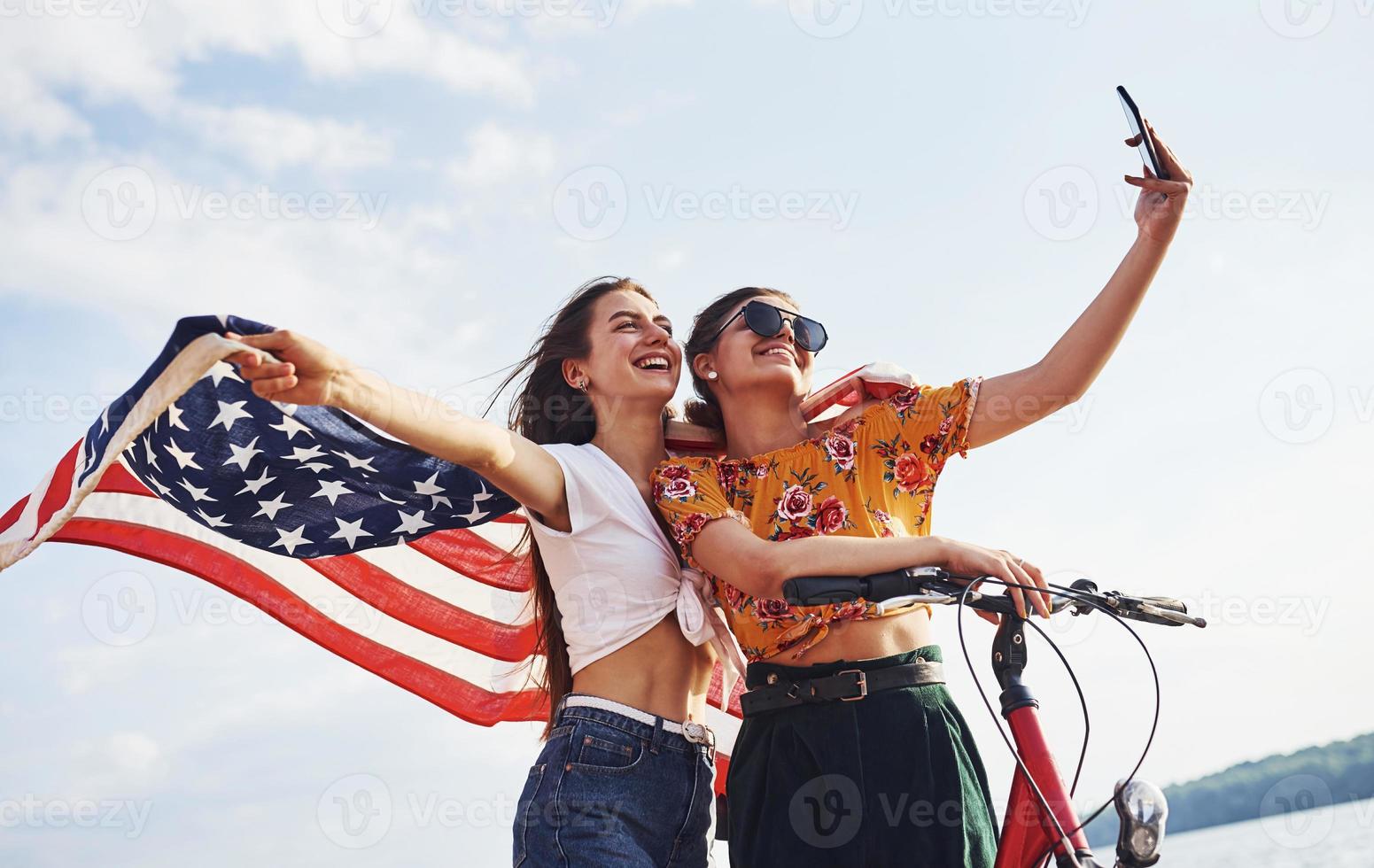 zwei patriotische fröhliche frauen mit fahrrad und usa-flagge in den händen machen selfie foto