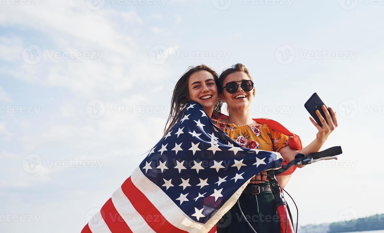 zwei patriotische fröhliche frauen mit fahrrad und usa-flagge in den händen machen selfie foto