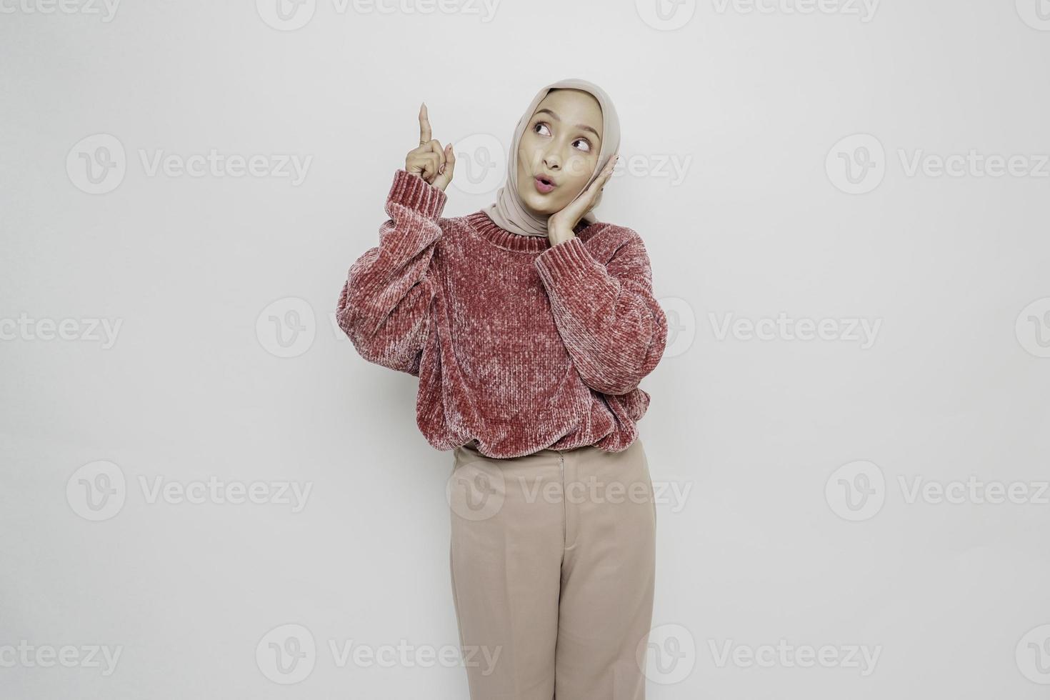 schockierte asiatische muslimische Frau mit rosa Pullover und Hijab, die auf den Kopierraum auf ihr zeigt, isoliert durch weißen Hintergrund foto