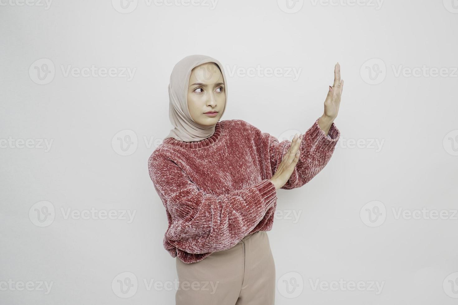 Schockierte asiatische muslimische Frau mit rosafarbenem Pullover und Hijab, die auf den Kopierbereich neben ihr zeigt, isoliert durch weißen Hintergrund foto