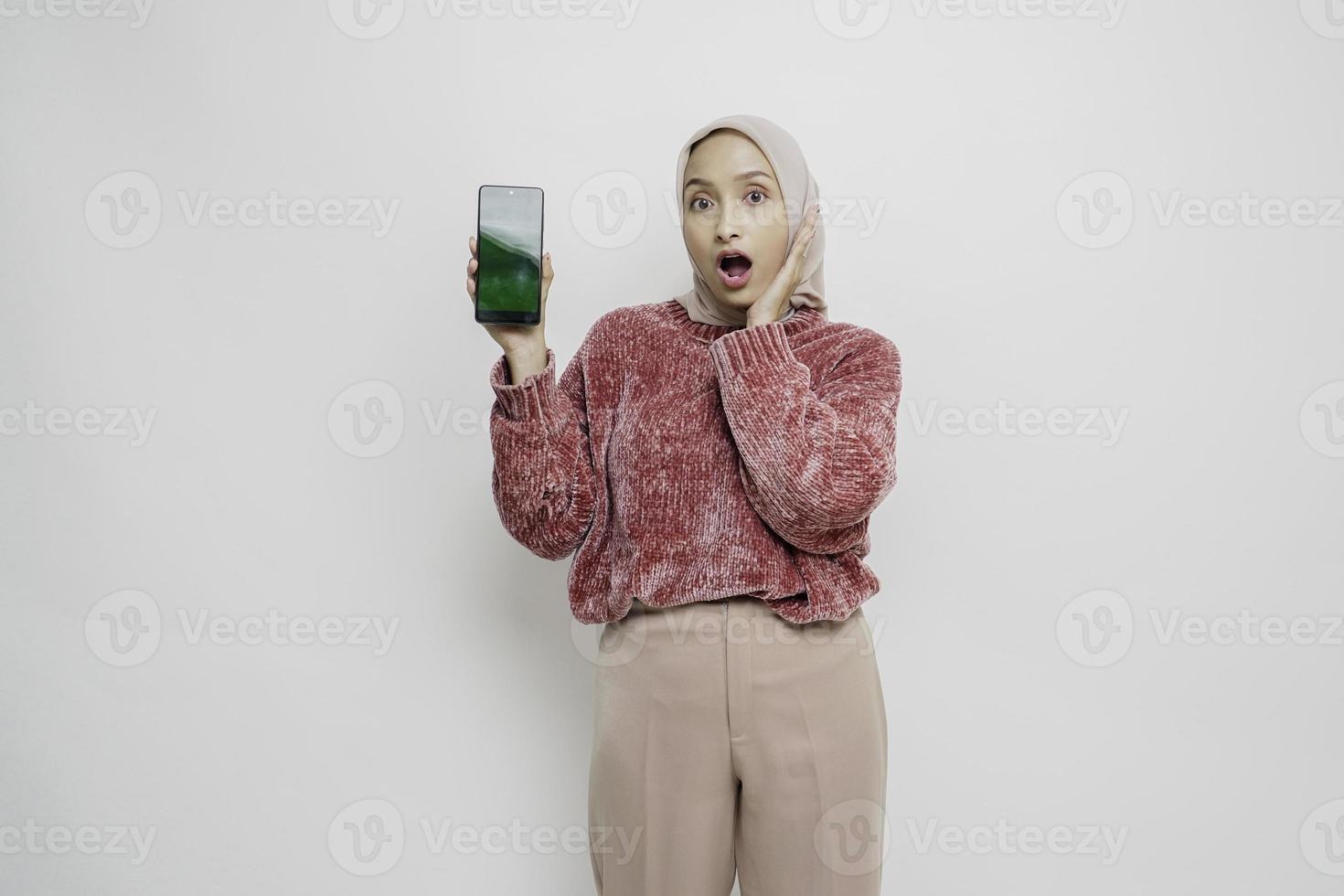 überraschte asiatische muslimische Frau mit rosa Pullover und Hijab, die auf ihrem Smartphone Platz für Kopien zeigt, isoliert durch weißen Hintergrund foto