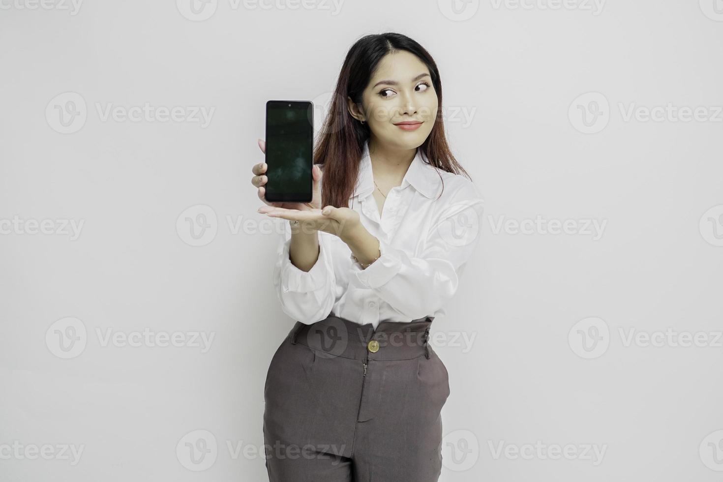 Aufgeregte asiatische Frau mit weißem Hemd, die auf den Kopierbereich neben ihr zeigt, während sie ihr Telefon hält, isoliert durch weißen Hintergrund foto