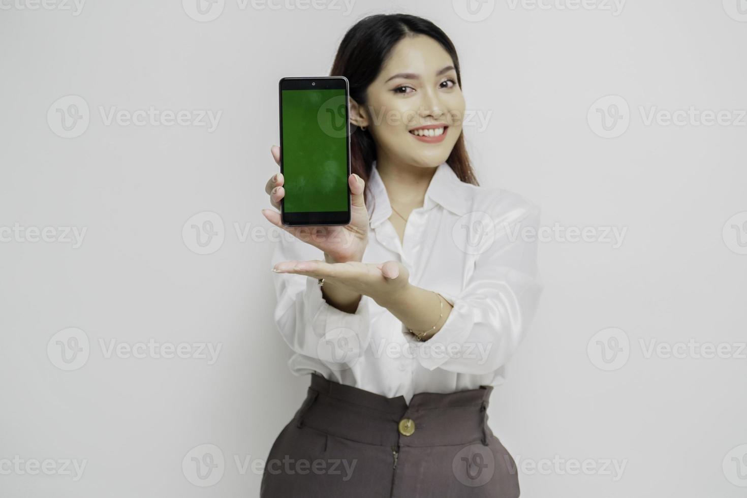 Aufgeregte asiatische Frau mit weißem Hemd, die auf den Kopierbereich neben ihr zeigt, während sie ihr Telefon hält, isoliert durch weißen Hintergrund foto