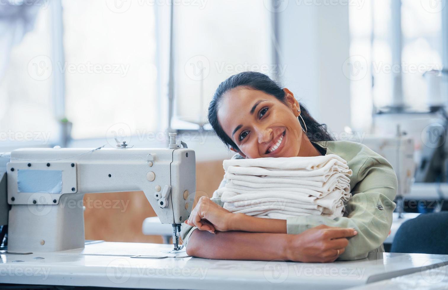 junge positive schneiderin sitzt mit kleidung am tisch foto