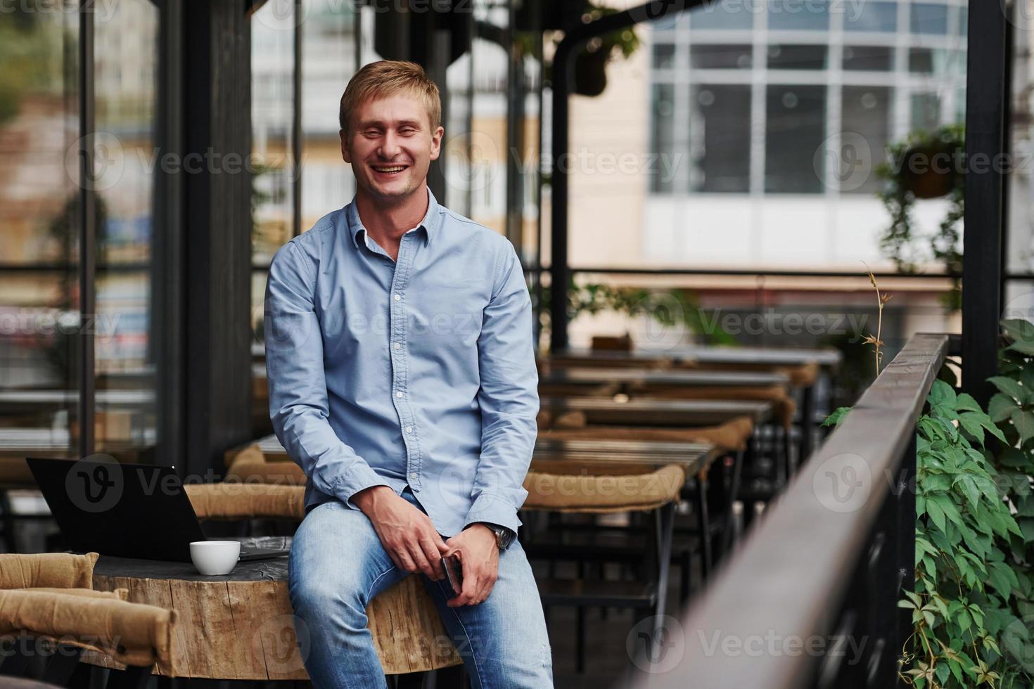 lächelnd für die Kamera. Vorderansicht des Typen, der tagsüber mit seinem Laptop in einem modernen Café ist foto