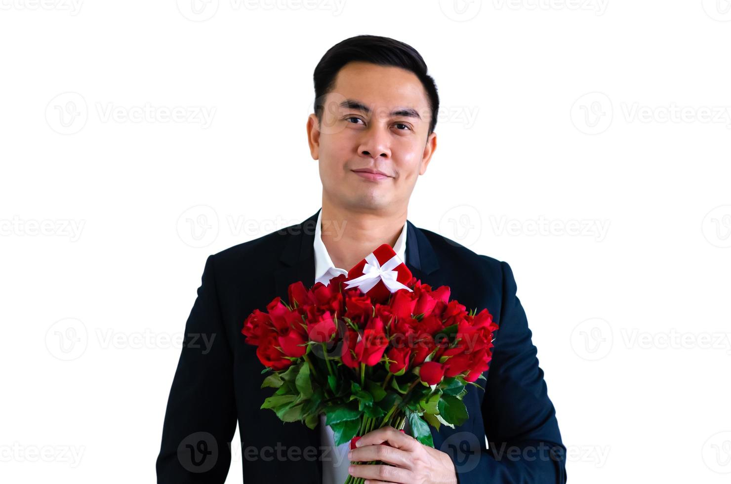 asiatischer mann im schwarzen anzug mit einem strauß roter rosen und einer roten geschenkbox, die auf weißem hintergrund für ein jubiläums- oder valentinstagkonzept isoliert sind. foto