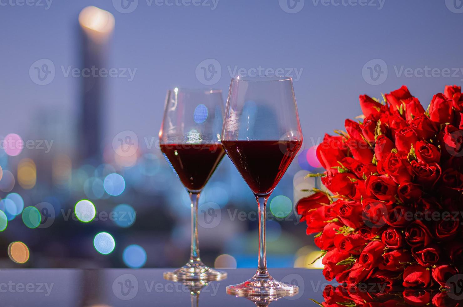 zwei gläser rotwein und roter rosenstrauß auf dem tisch mit bunten stadt-bokeh-lichtern zum jubiläum oder valentinstag-konzept. foto