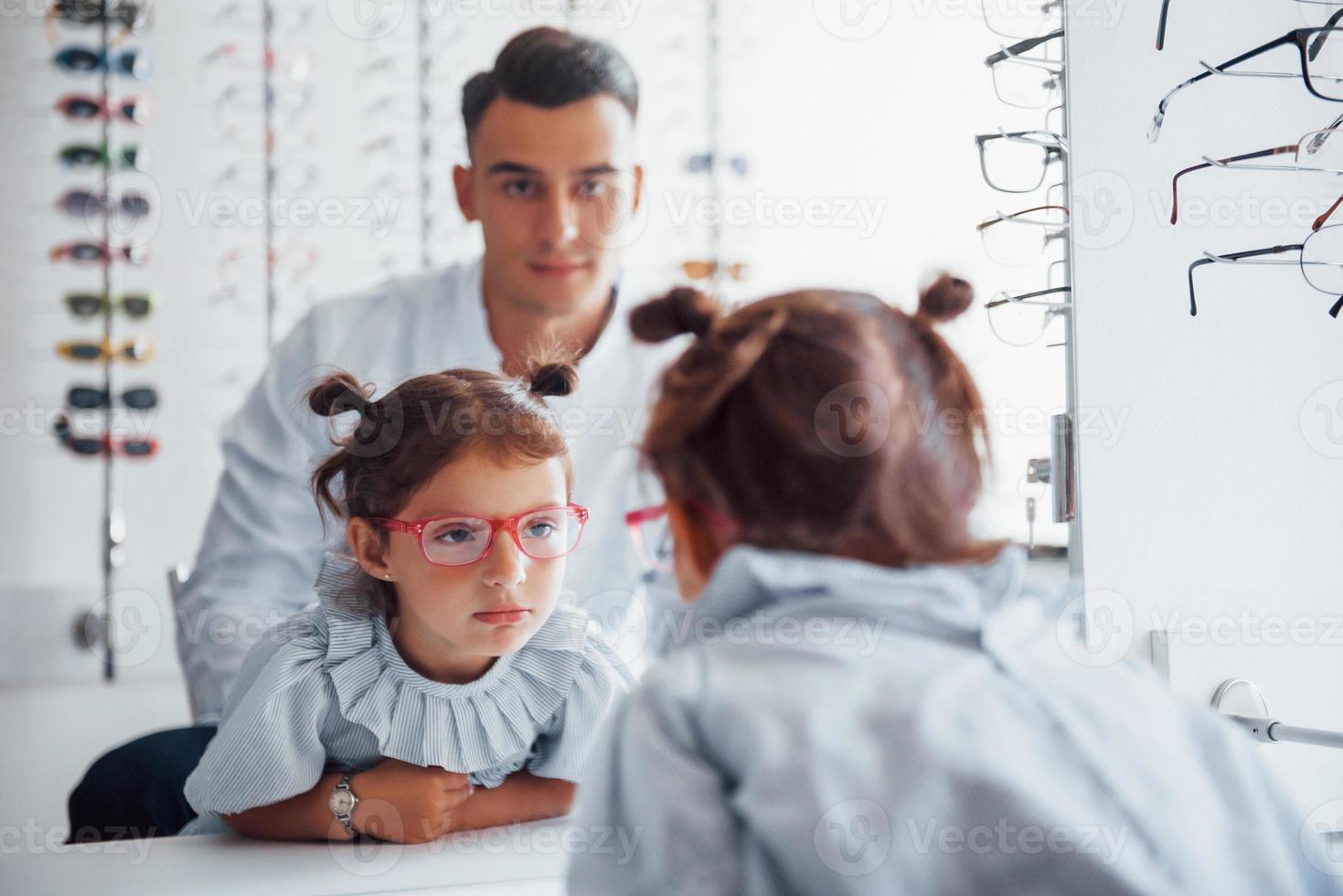 junger kinderarzt im weißen kittel hilft, neue brille für kleines mädchen zu bekommen foto