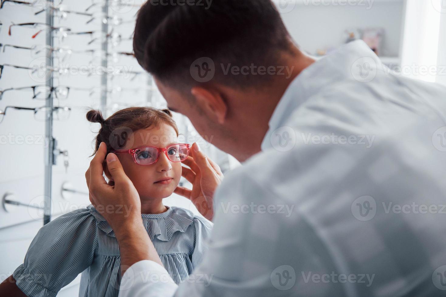 junger kinderarzt im weißen kittel hilft, neue brille für kleines mädchen zu bekommen foto