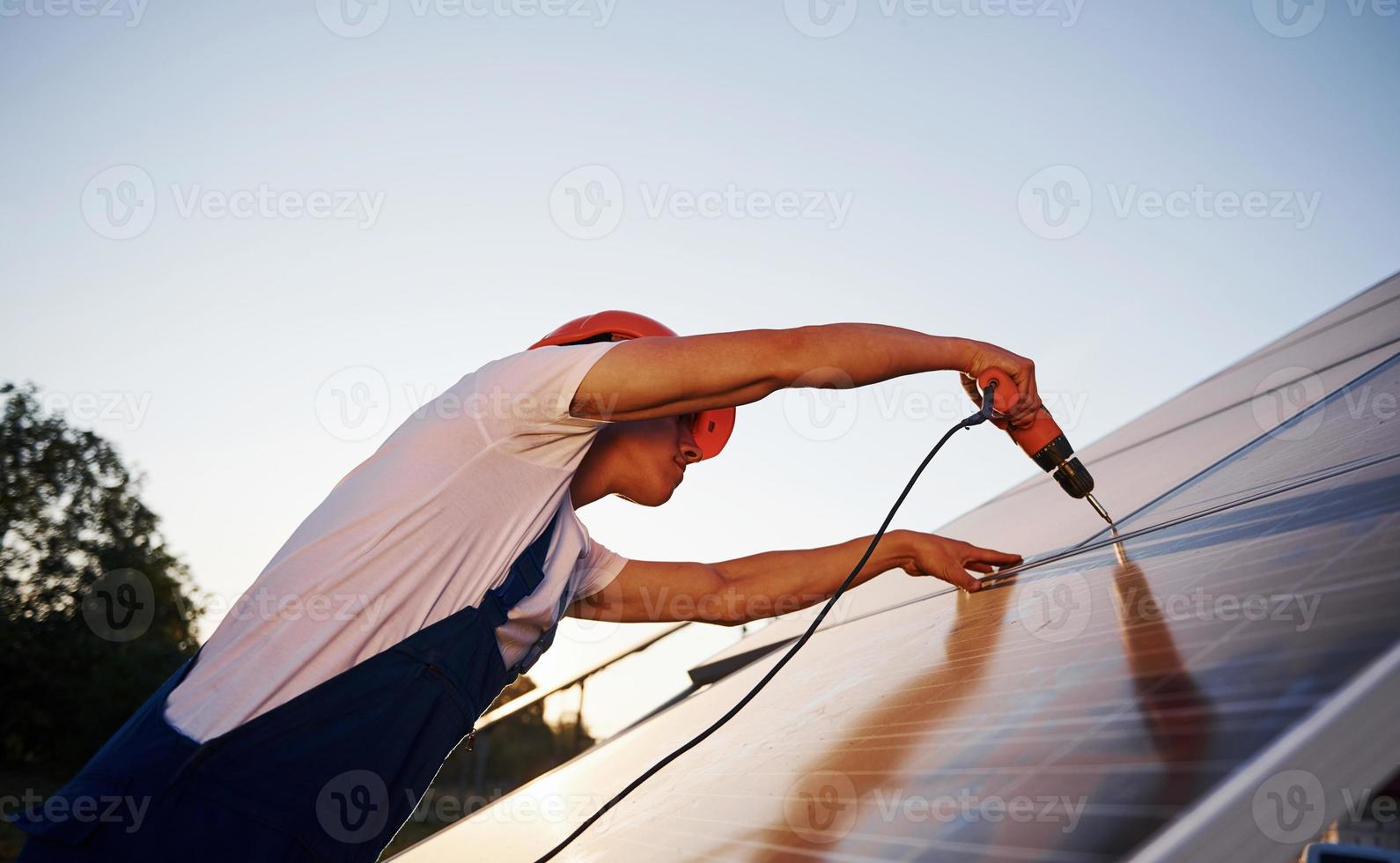 mit Akkuschrauber. männlicher Arbeiter in blauer Uniform im Freien mit Solarbatterien an sonnigen Tagen foto