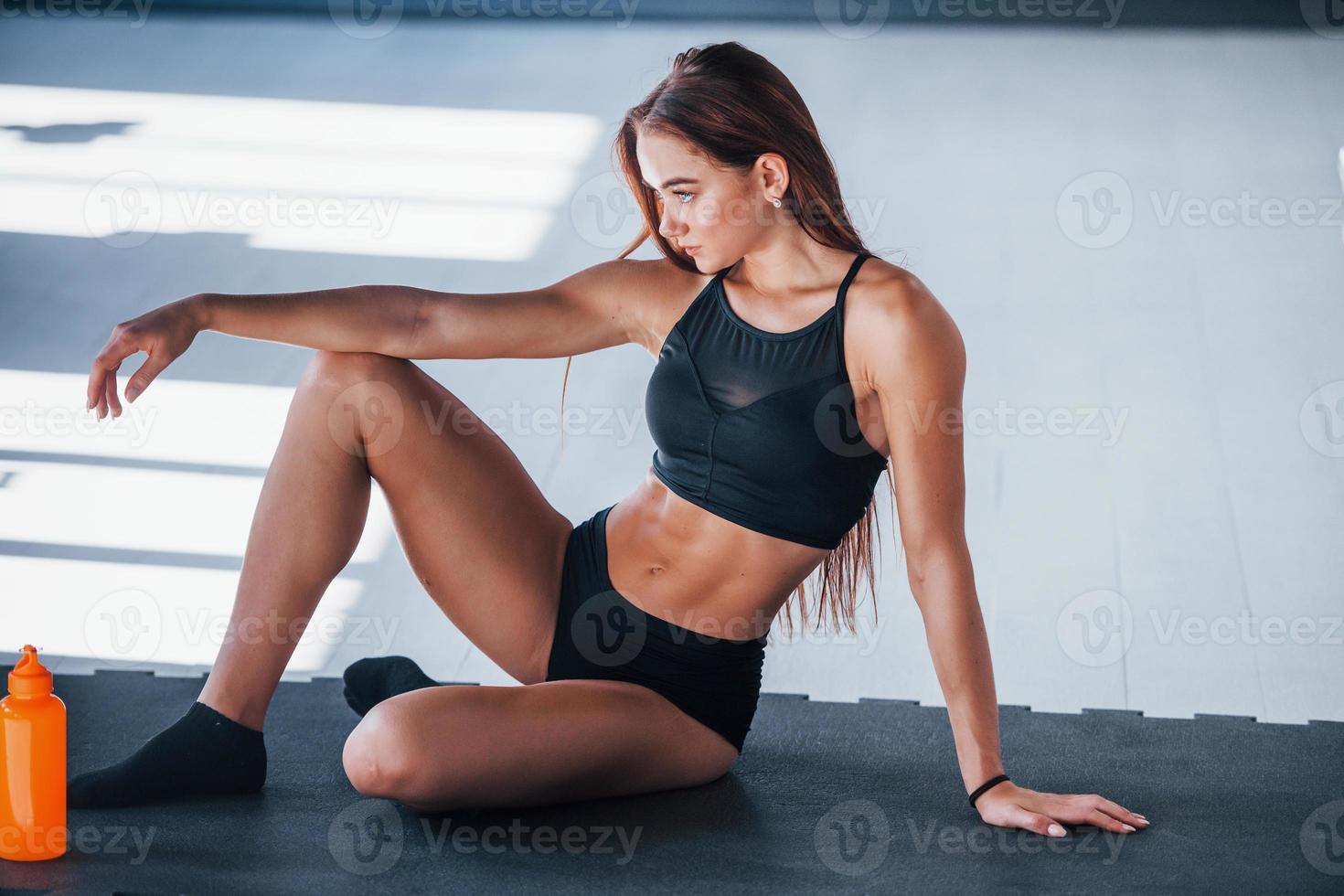 Junge Fitness-Frau mit schlankem Körpertyp sitzt mit einer Flasche Wasser auf der Matte foto