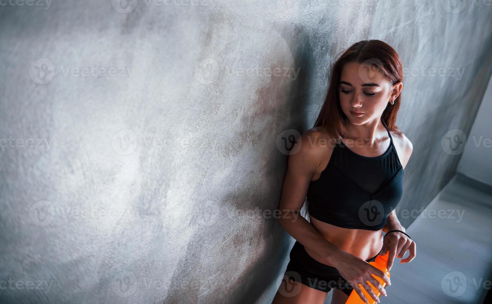 junge fitnessfrau mit schlankem körper und flasche wasser lehnt an der wand foto