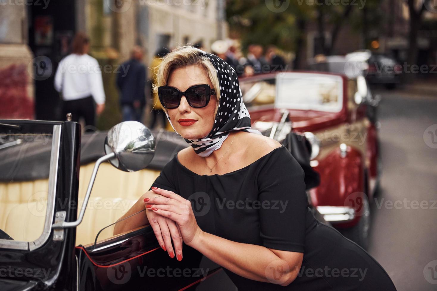 blonde Frau mit Sonnenbrille und in schwarzem Kleid, die sich an alte Oldtimer anlehnt foto