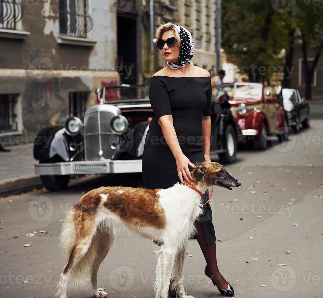 Blonde Frau mit Sonnenbrille und schwarzem Kleid in der Nähe eines alten Oldtimers mit ihrem Hund foto