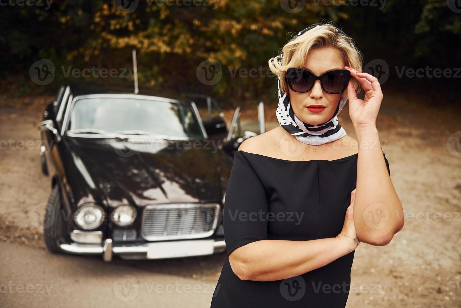 Blonde Frau mit Sonnenbrille und schwarzem Kleid in der Nähe eines alten Oldtimers foto