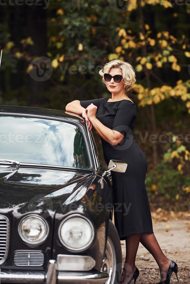 Blonde Frau mit Sonnenbrille und schwarzem Kleid in der Nähe eines alten Oldtimers foto