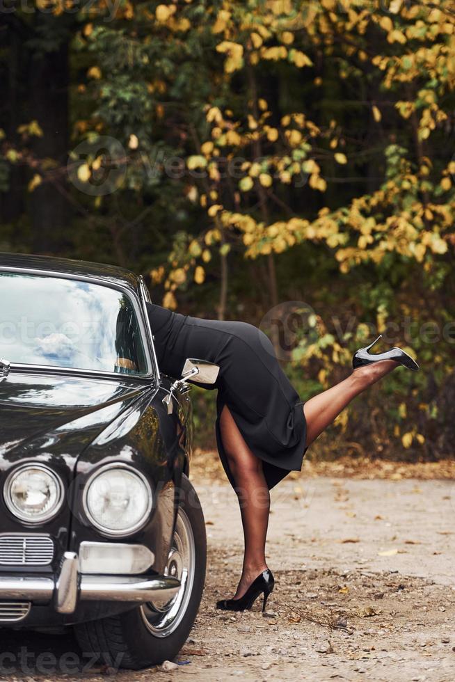 Frau auf High Heels und in schwarzem Kleid sieht mit erhobenem Bein in ein Retro-Auto foto