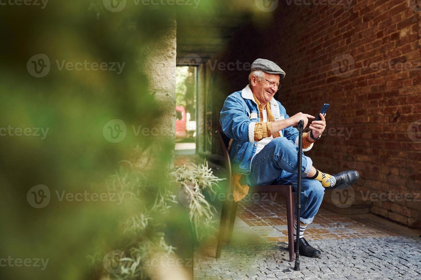 Stylischer Senior in modischer Kleidung und Brille sitzt auf dem Stuhl und telefoniert foto