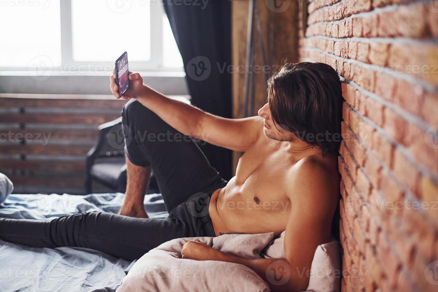 Ein hemdloser sexy Mann mit Telefon in der Hand ruht sich morgens allein in seinem Schlafzimmer aus foto