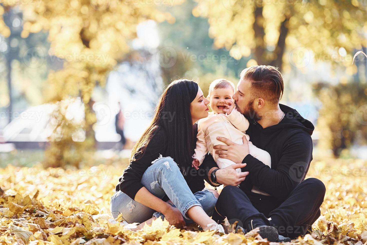 auf dem Boden sitzen. Fröhliche Familie, die sich zusammen mit ihrem Kind im schönen Herbstpark amüsiert foto