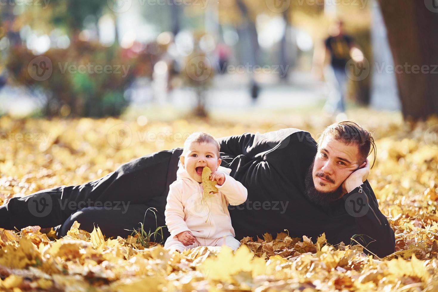 Vater in Freizeitkleidung, der mit seinem Kind auf dem Boden liegt, ist im schönen Herbstpark foto