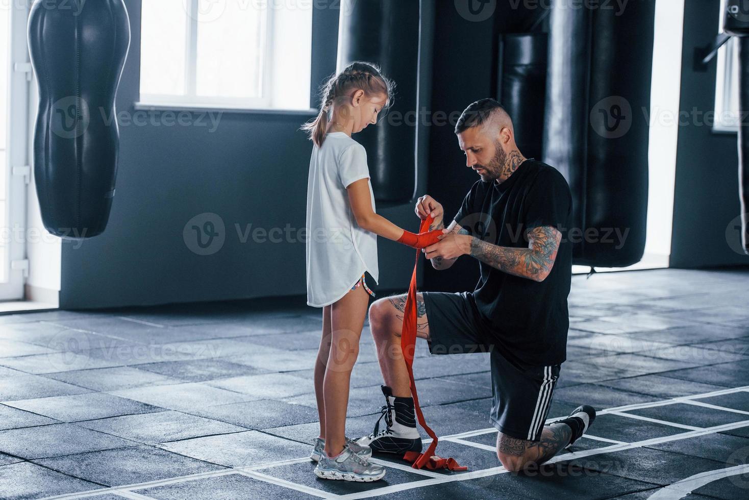 beim Tragen der Bandagen helfen. Der junge tätowierte Boxtrainer unterrichtet ein süßes kleines Mädchen im Fitnessstudio foto