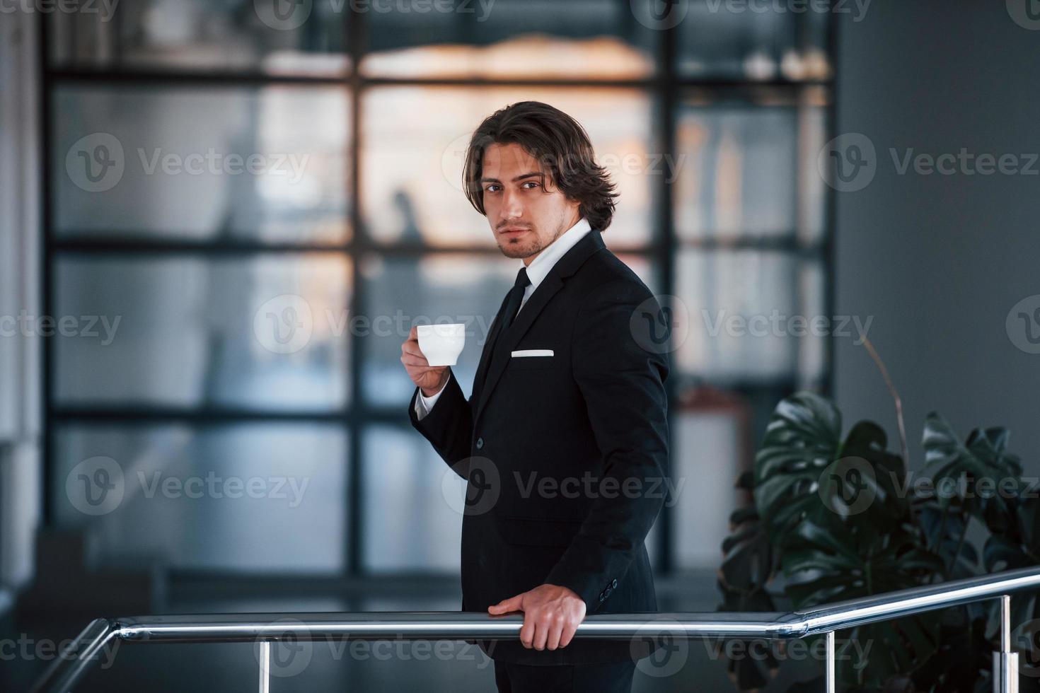 Porträt eines hübschen jungen Geschäftsmannes in schwarzem Anzug und Krawatte steht drinnen mit einer Tasse Getränk foto
