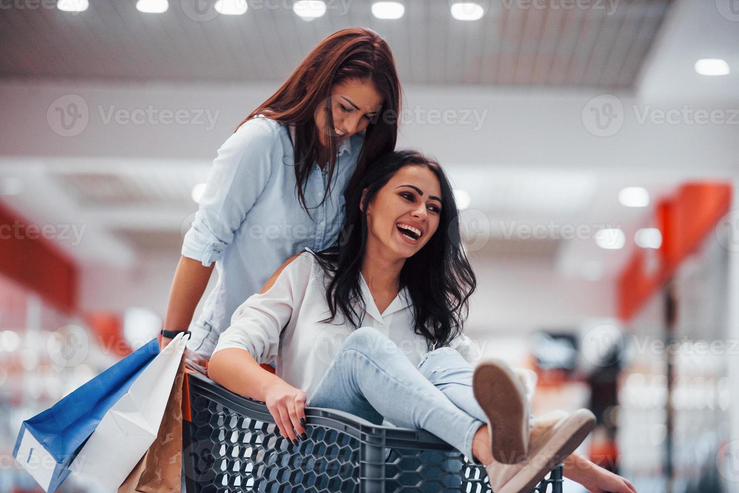 zwei junge frauen haben spaß beim rennen und reiten im einkaufskorb im supermarkt foto