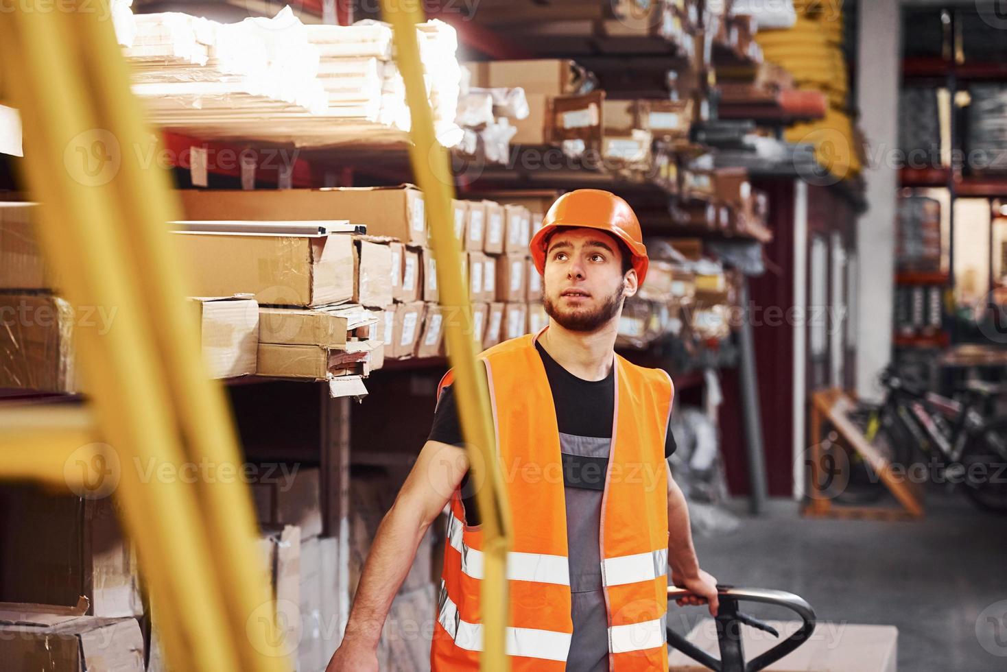 junger männlicher arbeiter in uniform ist im lager und schiebt hubwagen foto