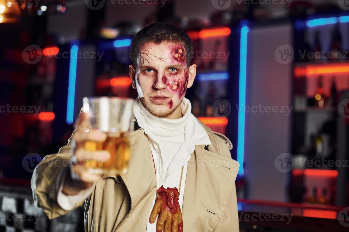 mit Getränk in der Hand. Porträt des Mannes, der auf der thematischen Halloween-Party in Zombie-Make-up und Kostüm ist foto