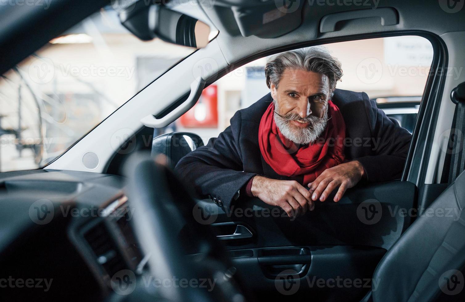Modischer alter Mann mit grauem Haar und Schnurrbart, der drinnen im Salon nach einem neuen Auto sucht foto