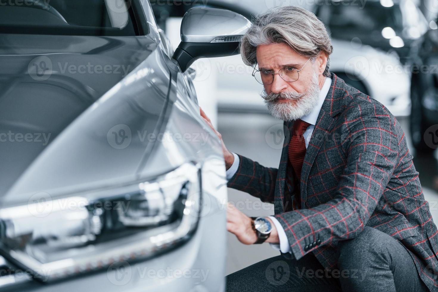 Modischer alter Mann mit grauem Haar und Schnurrbart, der drinnen im Salon ein neues Auto berührt foto