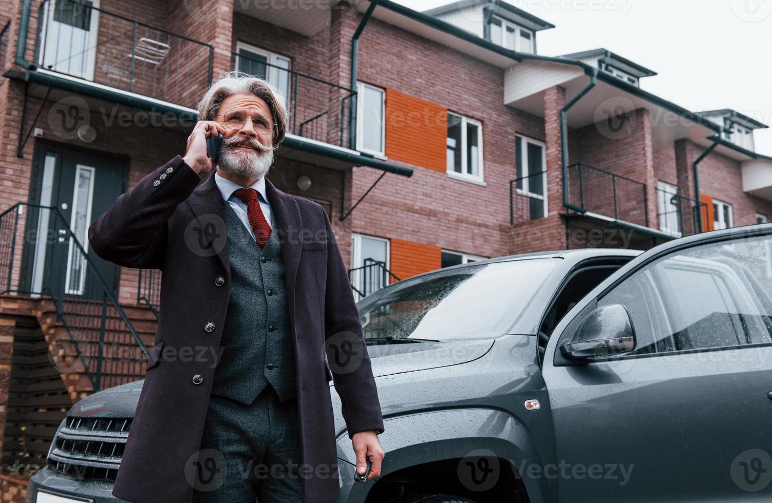 Modischer Senior mit grauem Haar und Bart telefoniert draußen auf der Straße in der Nähe seines Autos foto