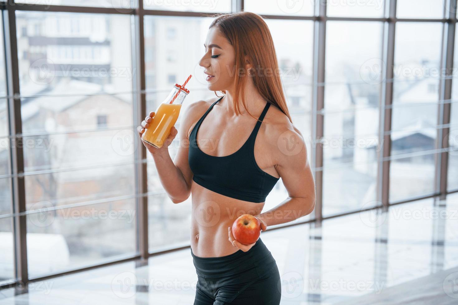 sportliche frau mit schönem körper steht drinnen mit einer flasche orangensaft und apfel in den händen foto