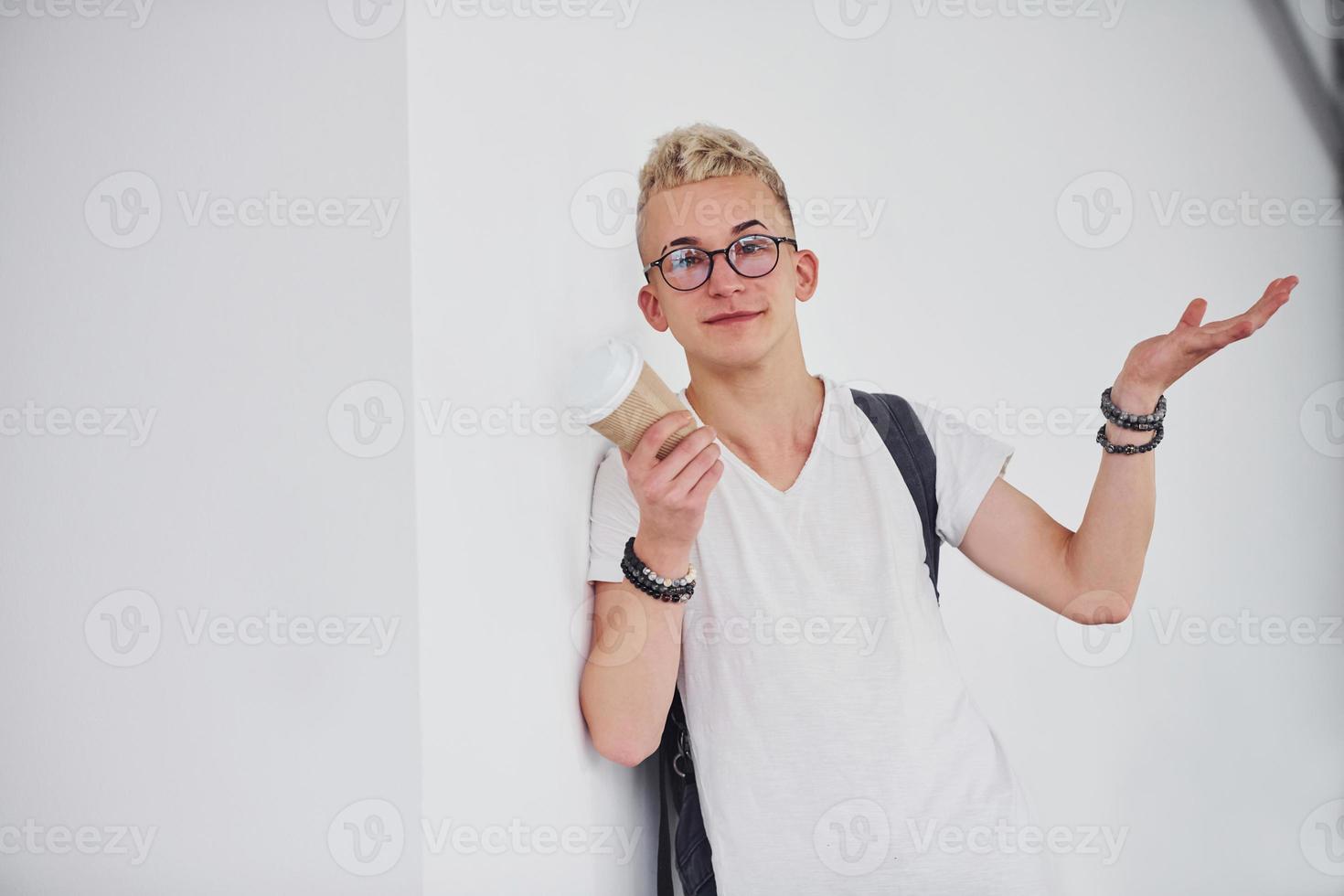 student in lässiger kleidung und mit rucksack steht drinnen gegen eine weiße wand mit einer tasse getränk foto