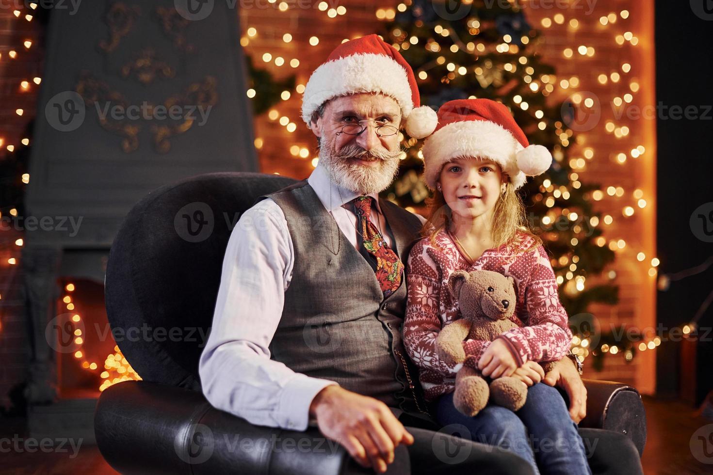 Fröhlicher älterer Mann mit grauem Haar und Bart sitzt mit kleinem Mädchen in Weihnachtsmützen und Teddybär foto