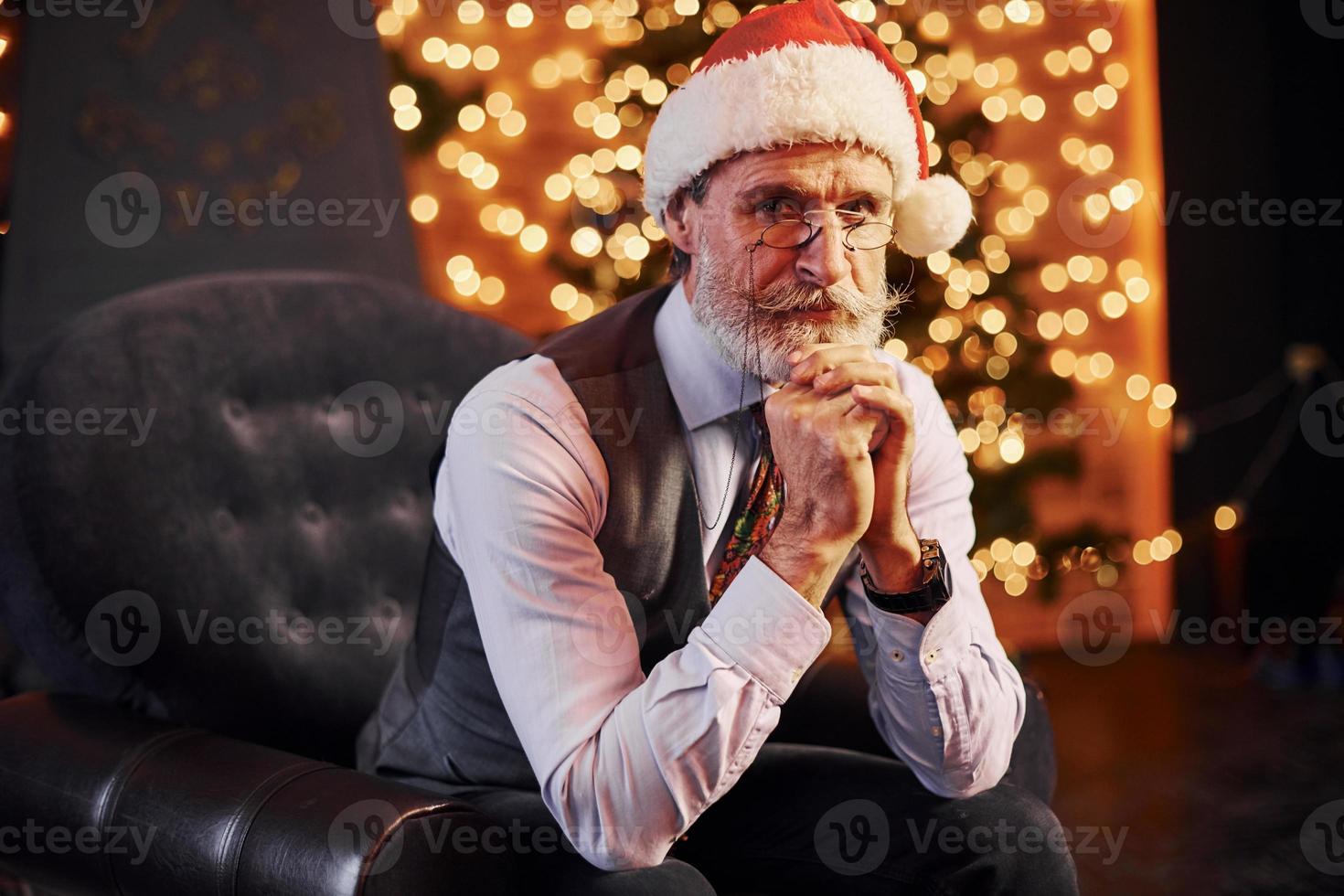 porträt eines stilvollen seniors mit grauem haar und bart, der in einem dekorierten raum und in einer weihnachtsmütze sitzt foto