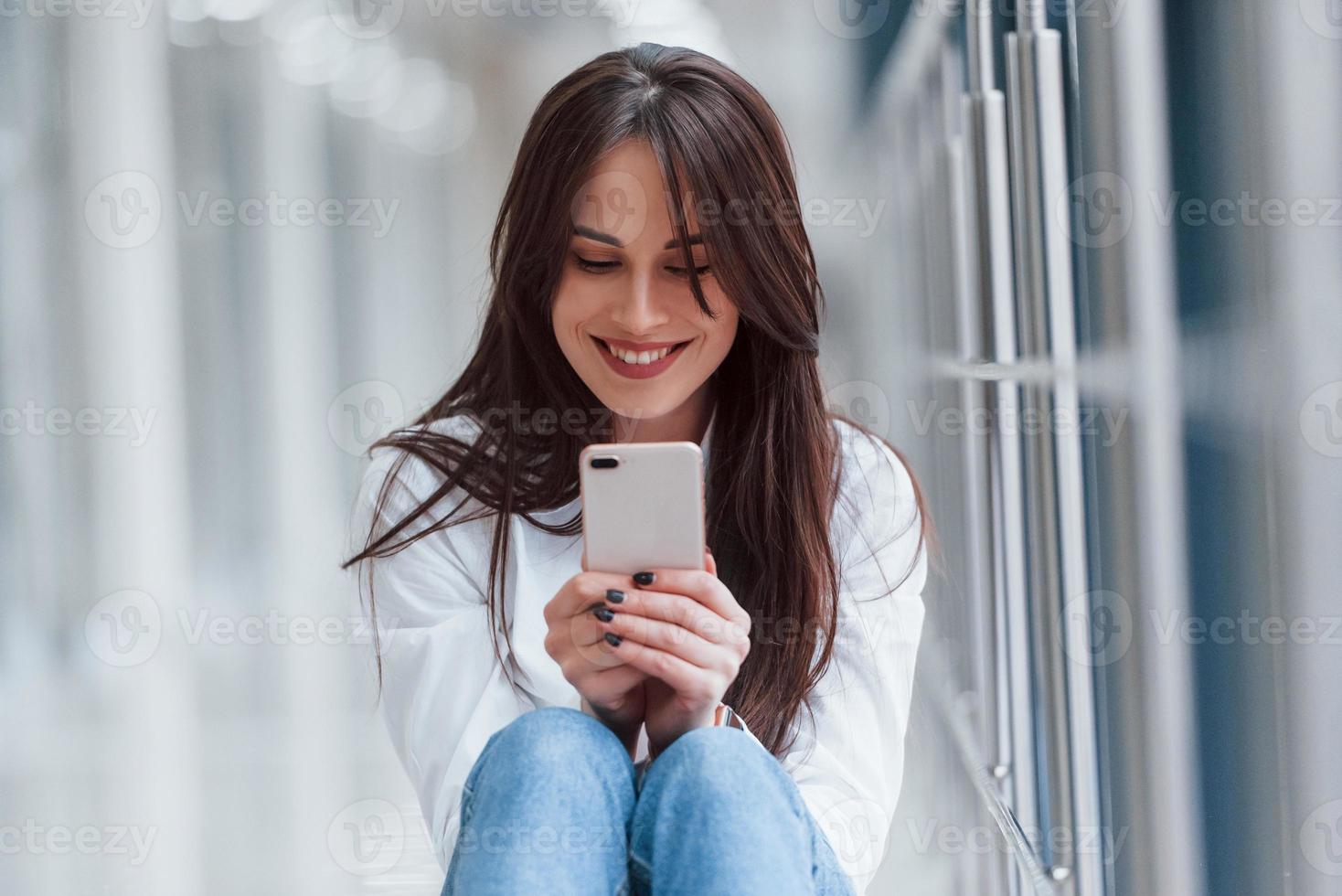 brünette in weißem hemd mit telefon in den händen sitzt tagsüber drinnen im modernen flughafen oder flur foto